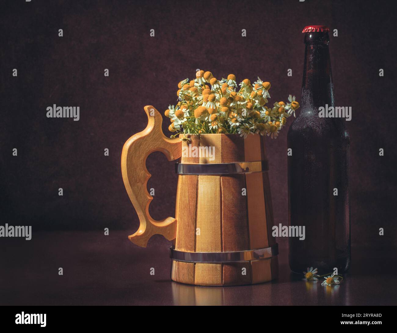 Still life with a bottle of beer with a wooden mug and camomiles imitating foam on a dark background Stock Photo