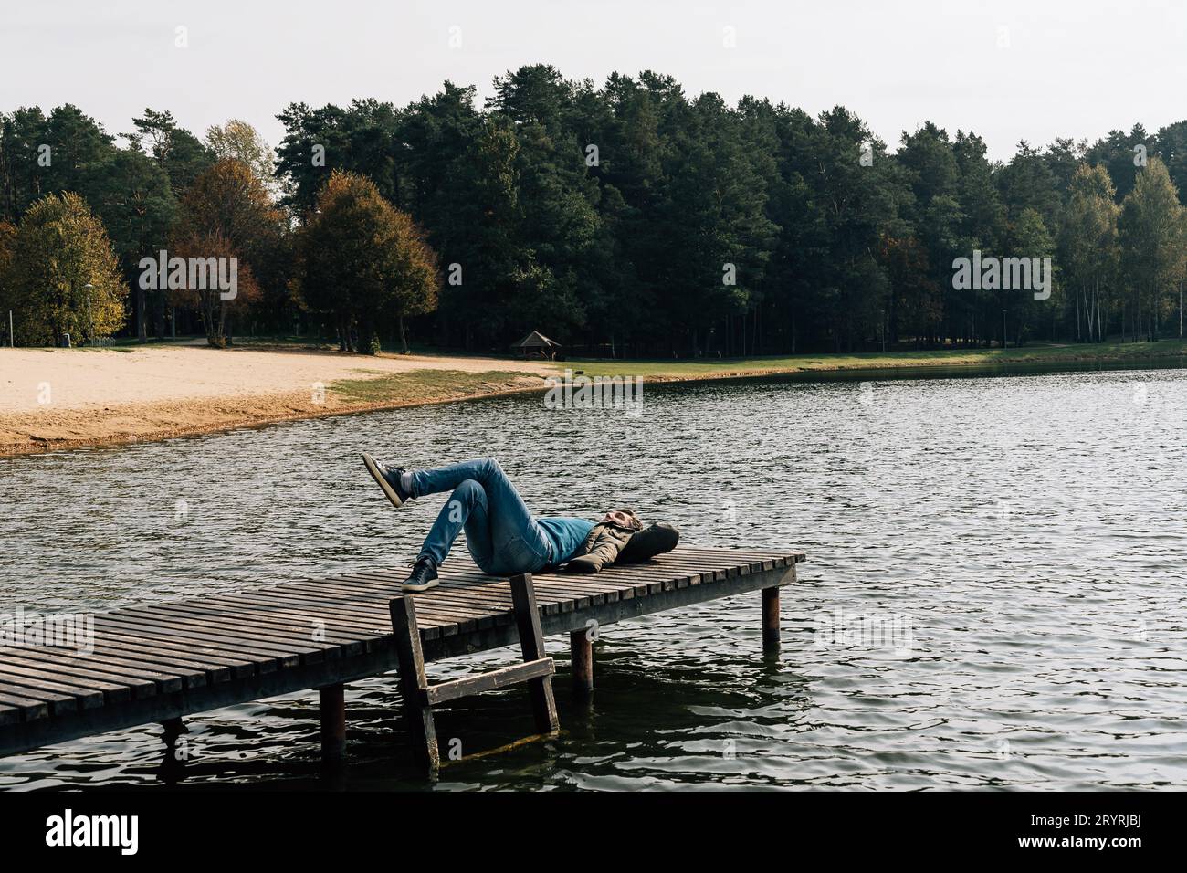 A man rests lying on a wooden pier on a lake in jeans and a jacket on an autumn day Stock Photo