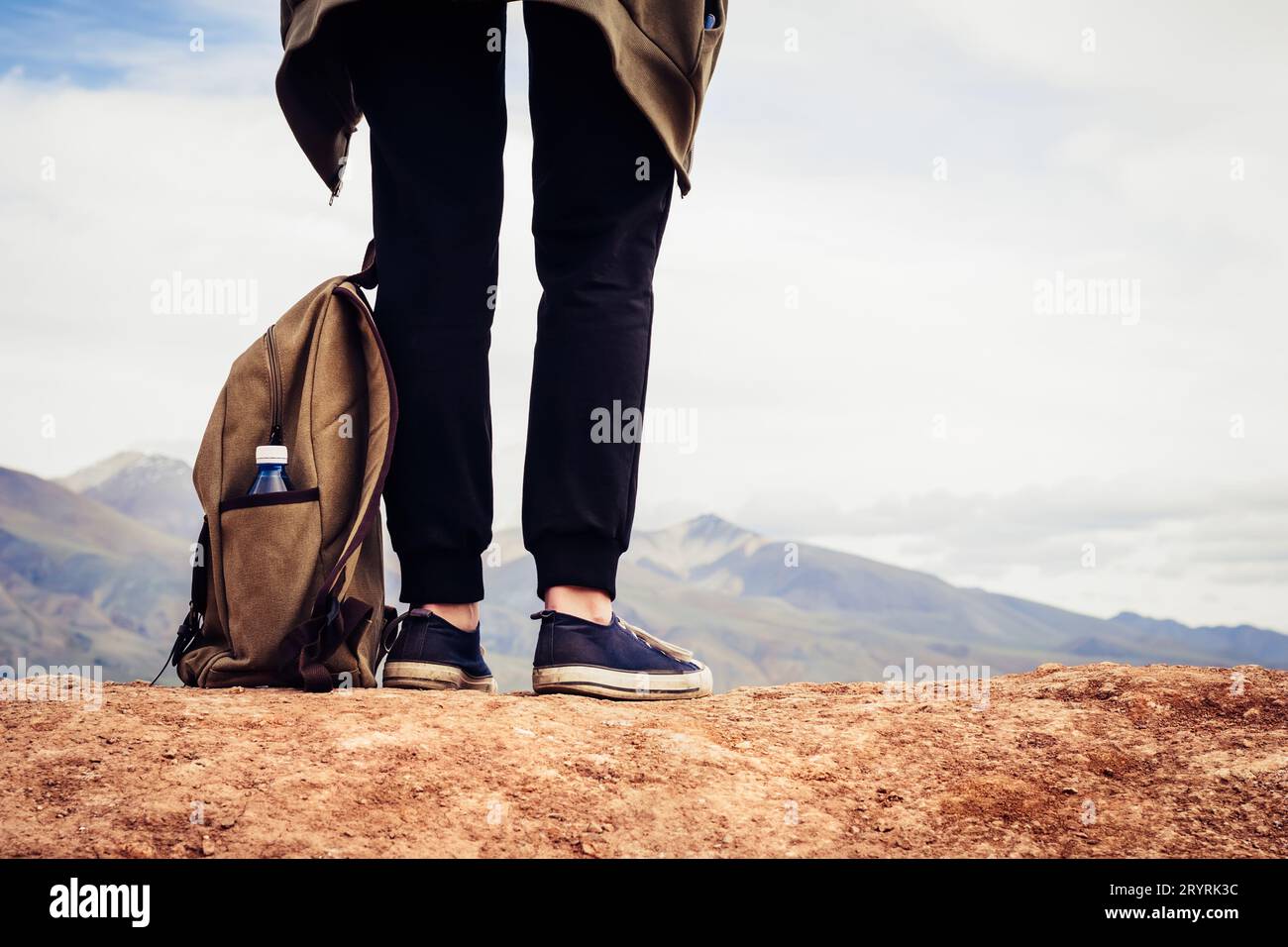 Girl tourist in black sports pants stands on hill. Legs of traveler and backpack with bottle of water close-up Stock Photo