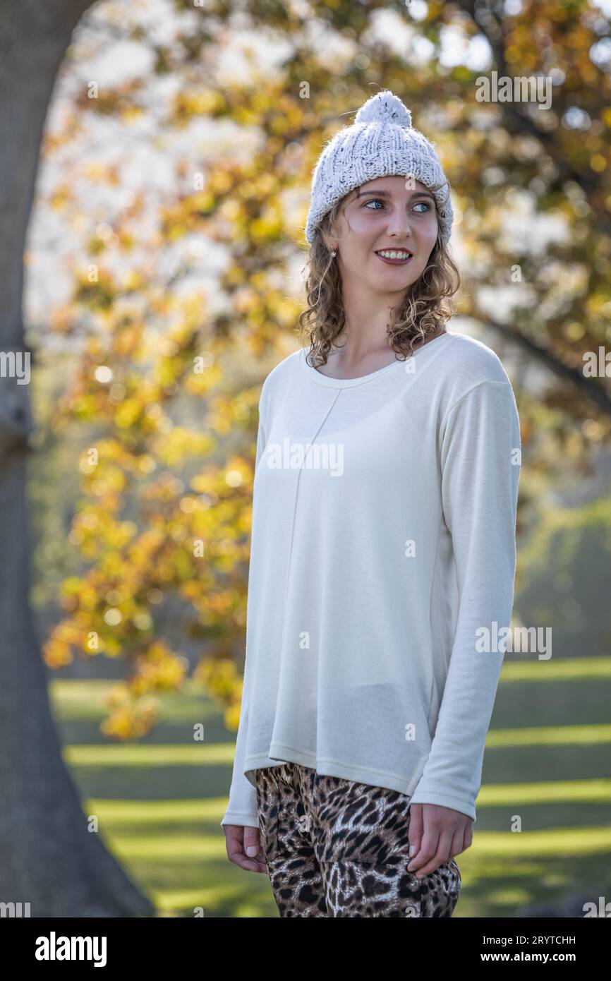 A young woman in a cream-colored sweater and a woolen hat gazes over her shoulder beneath the grand canopy of autumn trees Stock Photo