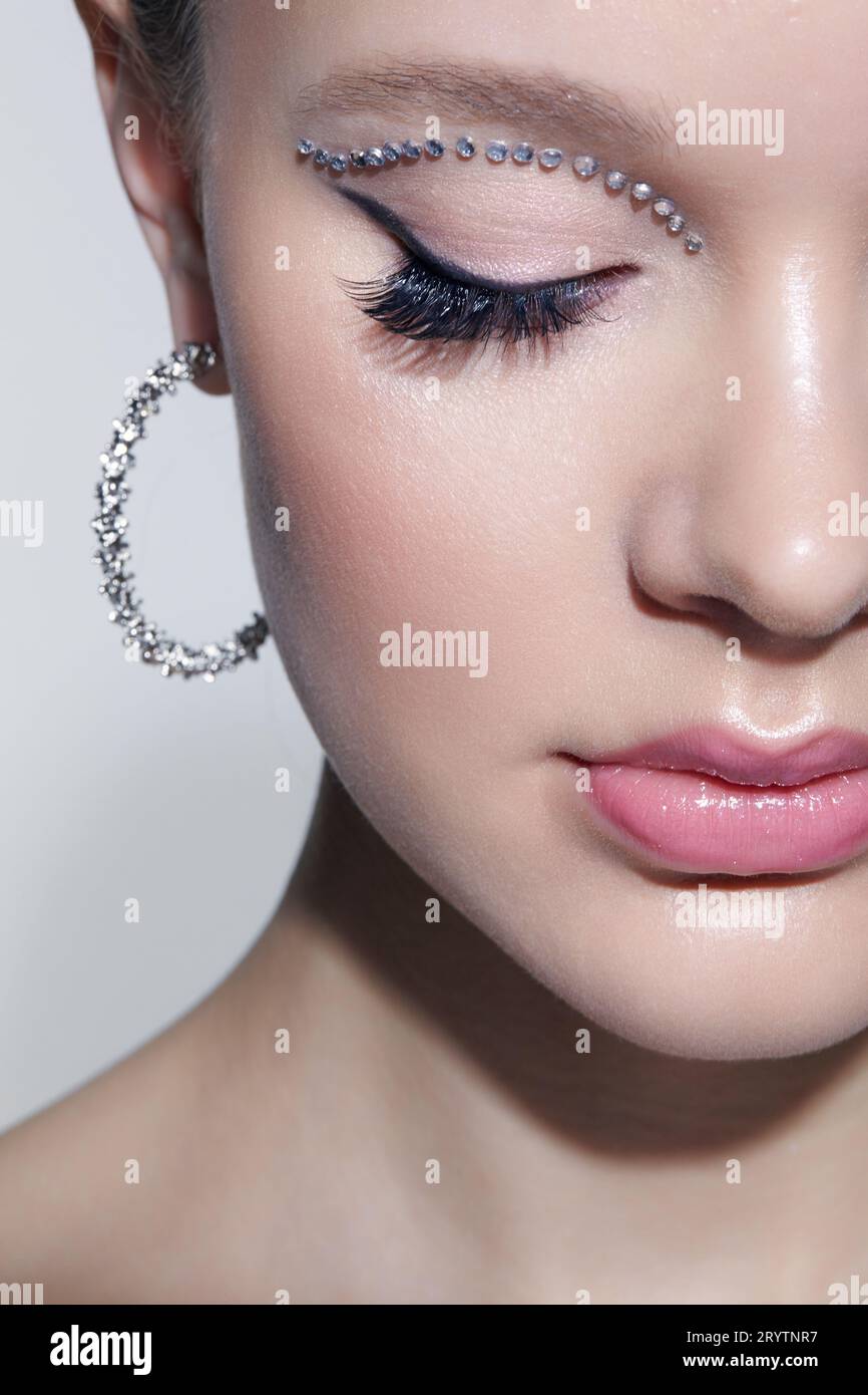 Closeup shot of human female face with unusual rhinestones makeup. Woman with closed eyes and earrings in the form of a shiny ri Stock Photo