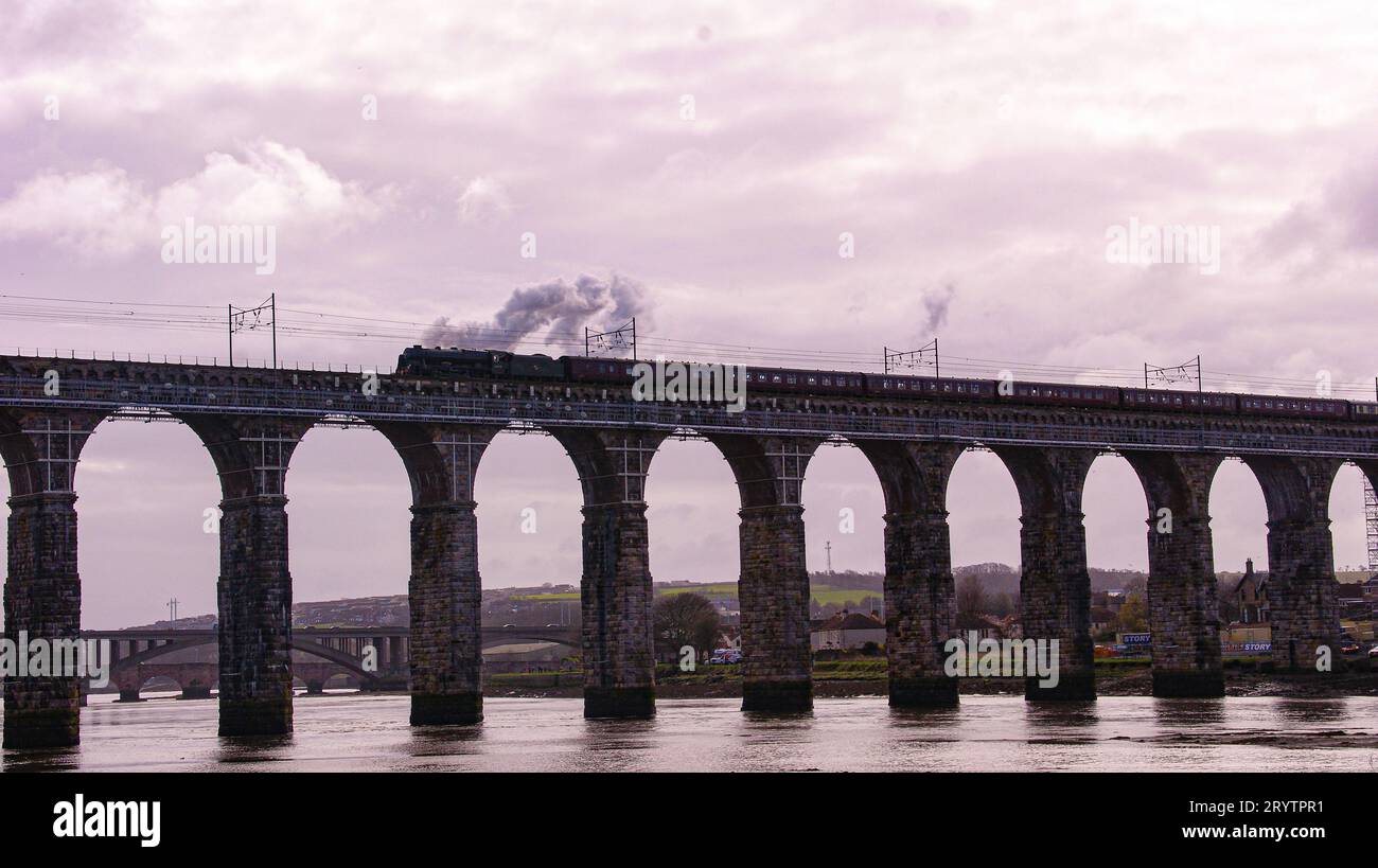 A train passing over a bridge, the river below and a town with hills in the background Stock Photo