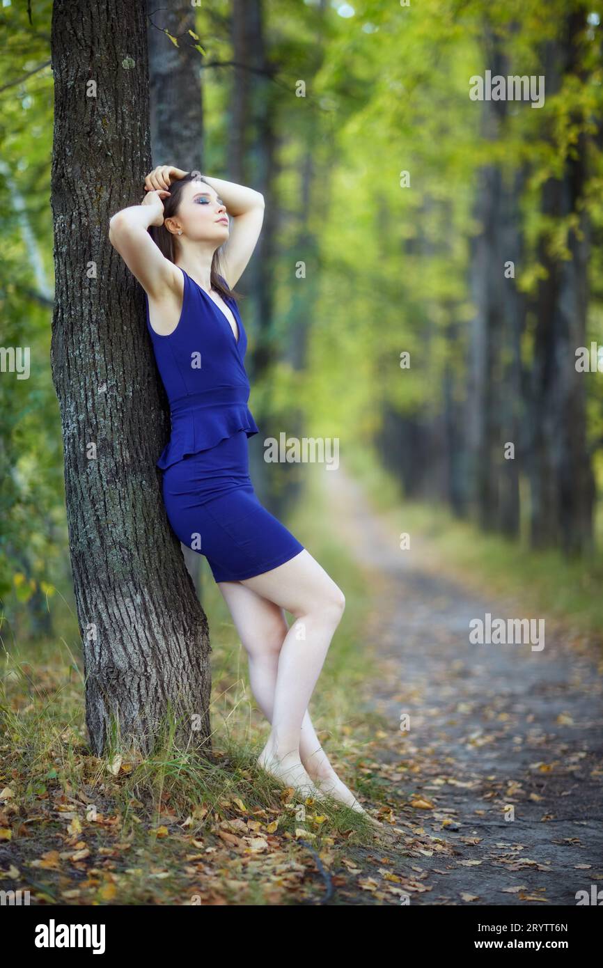 Female in blue dress leaning against a tree trunk in autumn linden tree alley. Stock Photo