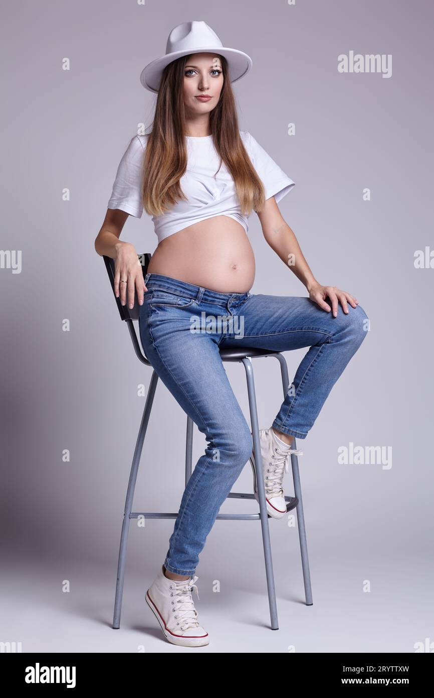 Young pretty pregnant woman in white t-shirt, and jeans sit on bar stool. Female with belly exposed. 5th month of pregnancy Stock Photo