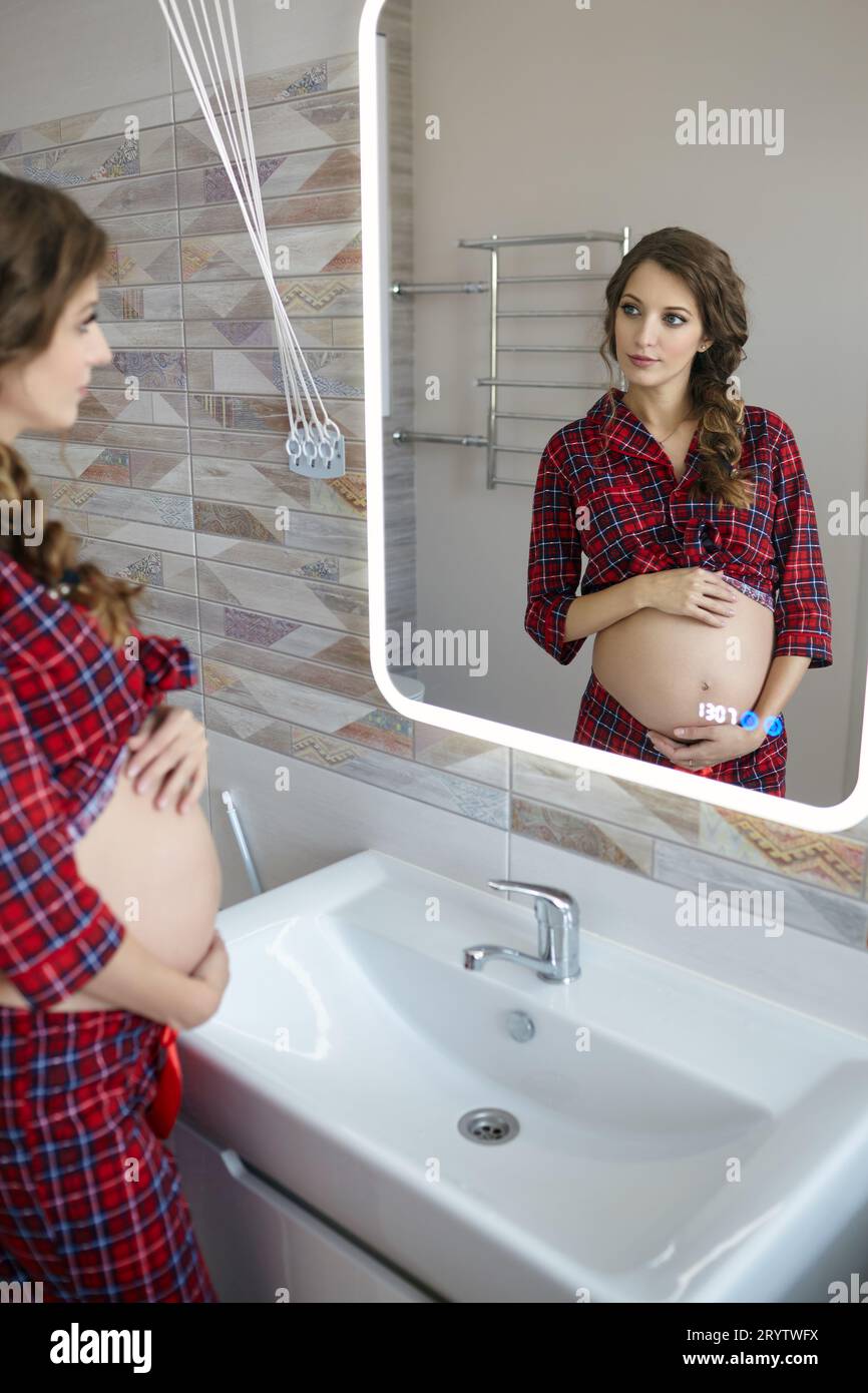 A pregnant woman in the bathroom looking in the mirror. Female dressed ...