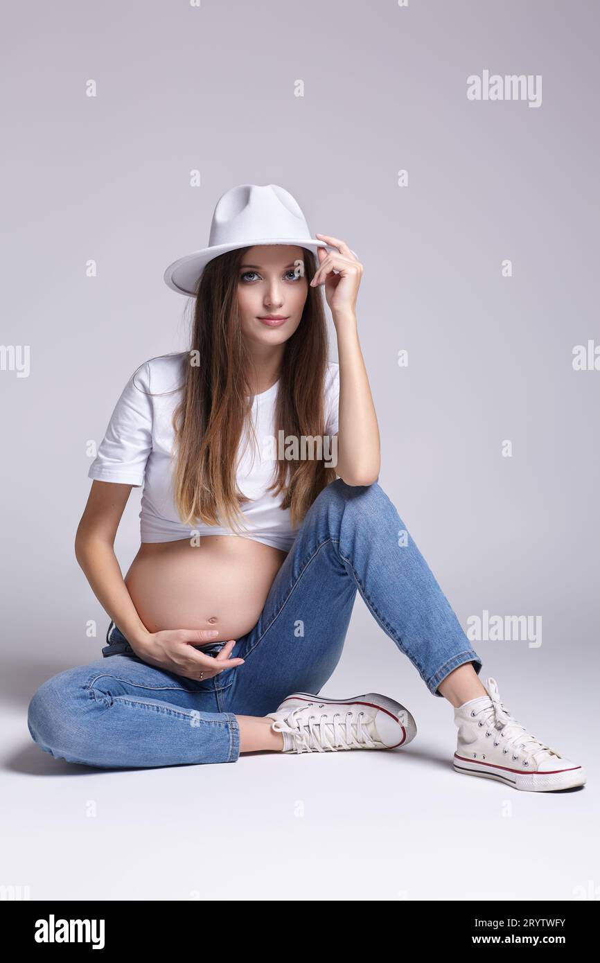 Young pretty pregnant woman in white t-shirt, hat and jeans sit on the floor on background. Female with belly exposed. 5th month Stock Photo