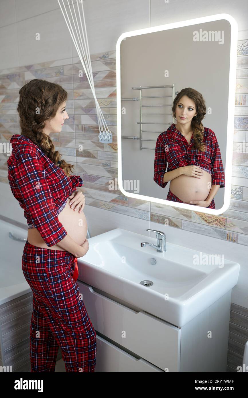 A pregnant woman in the bathroom looking in the mirror. Female dressed in red pajama and open belly enjoying her pregnancy state Stock Photo