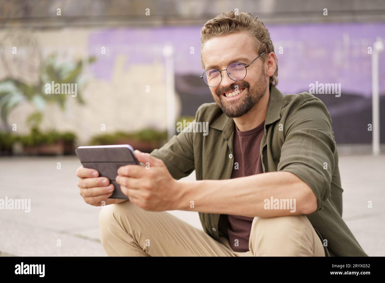 Mid-aged, handsome man sitting on the stairs of an embankment, engrossed in reading good news on his tablet PC. A genuine smile Stock Photo