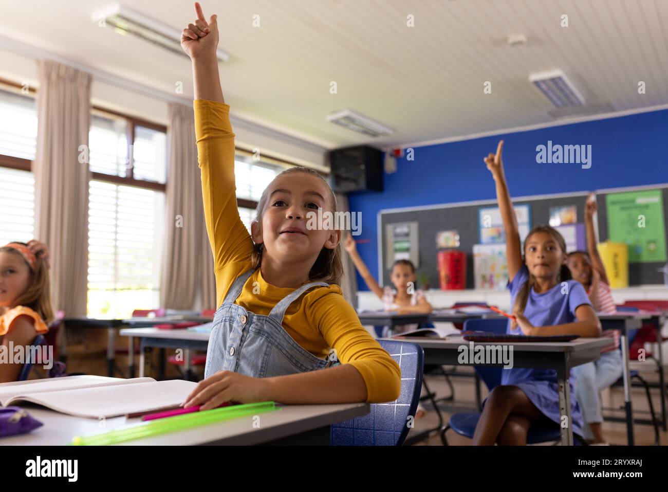 Happy diverse schoolgirls sitting at desks and raising hands in ...