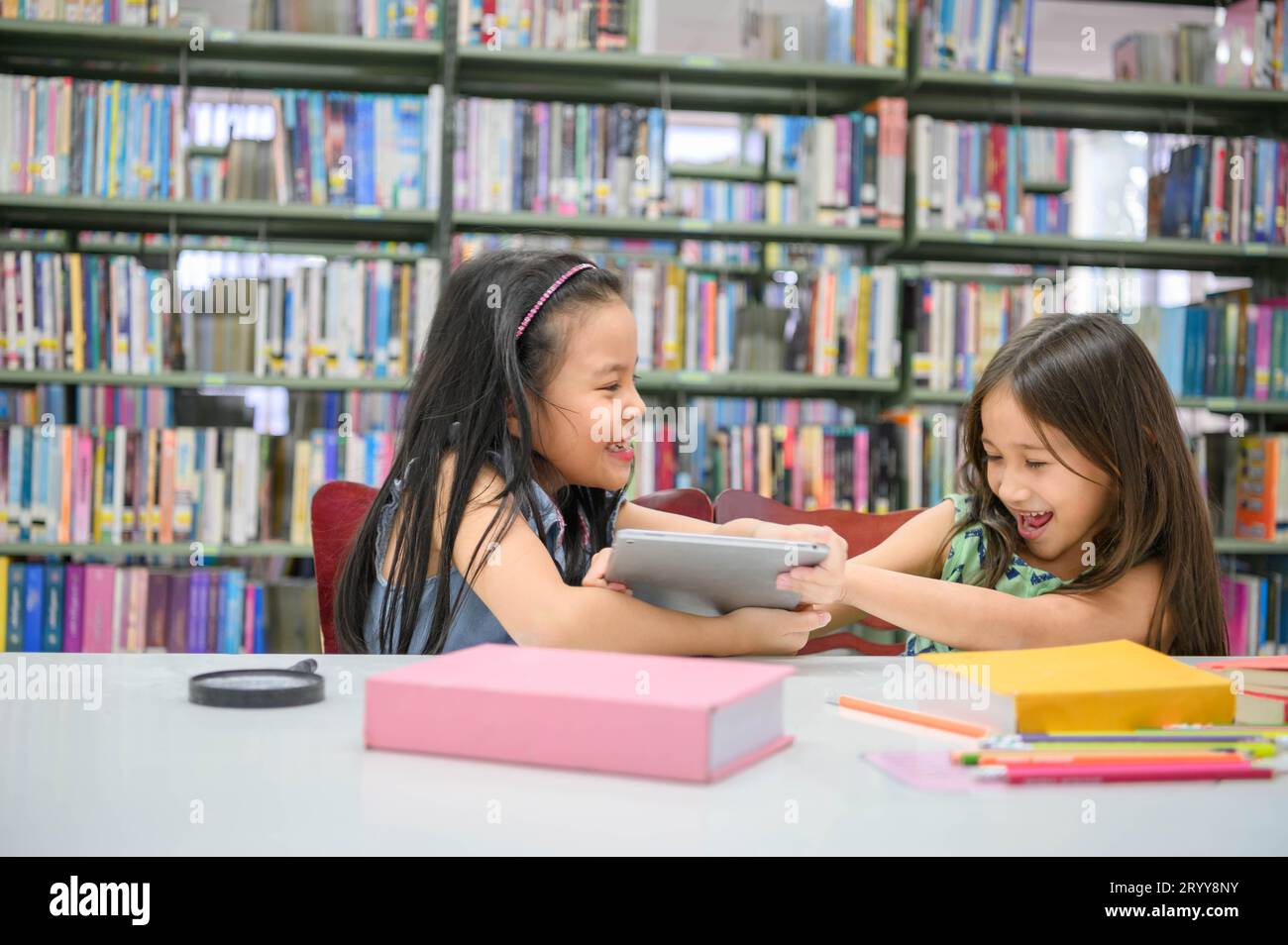Two girls fighting for tablet in the classroom while reading books. People lifestyles and education. Young friendship and Kids r Stock Photo