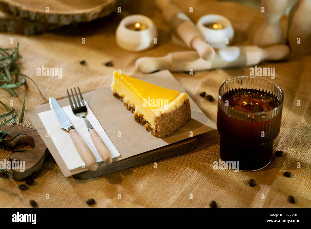 Alongside a slice of orange cheesecake, an iced Americano in a glass sits on a wooden board on a burlap tablecloth, complemented by the warm glow of b Stock Photo