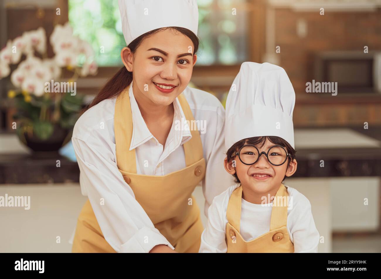 Portrait cute little Asian boy chef with eyeglasses and his mother looking to camera in home cooking kitchen happily and funny. Stock Photo