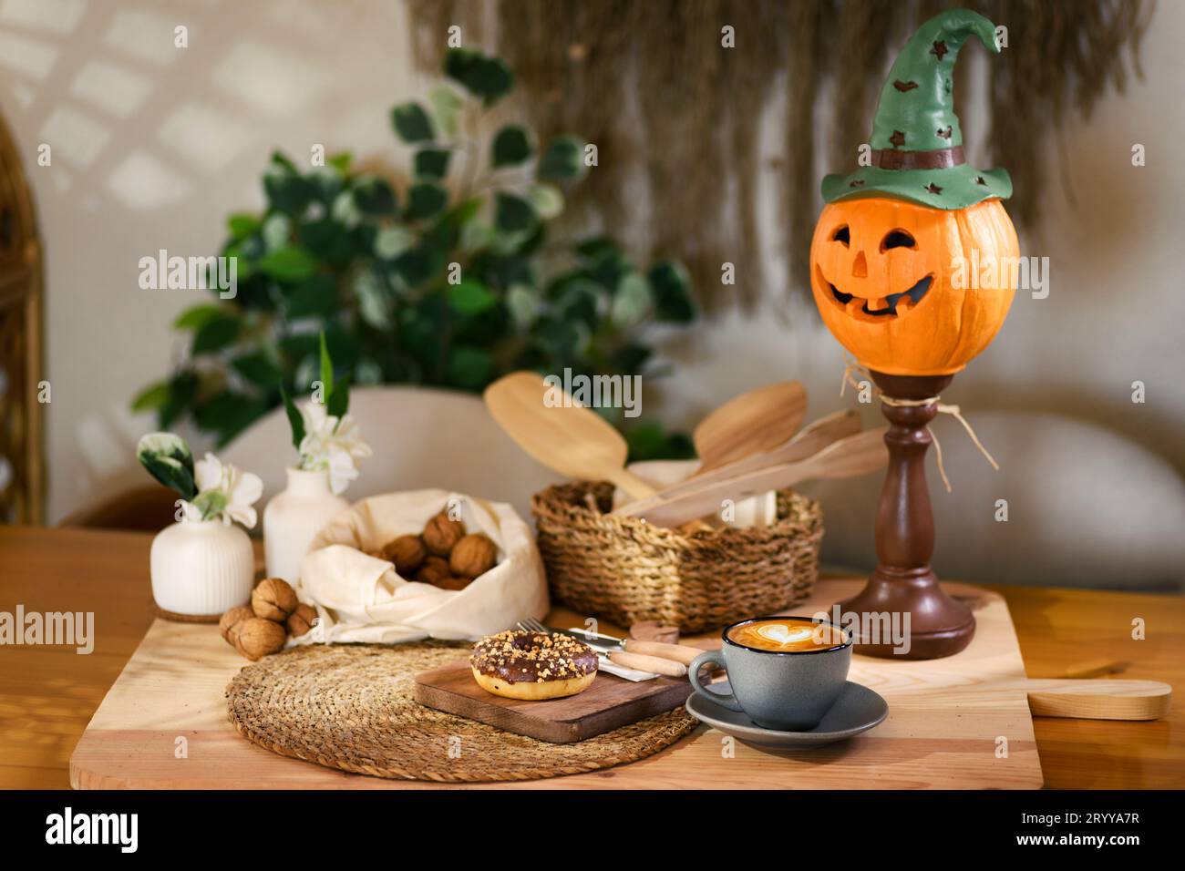 A single donut rests on a wooden plate, with walnuts on a white cloth behind. In a wicker basket, wooden kitchen tools lay, alongside a latte and a ja Stock Photo