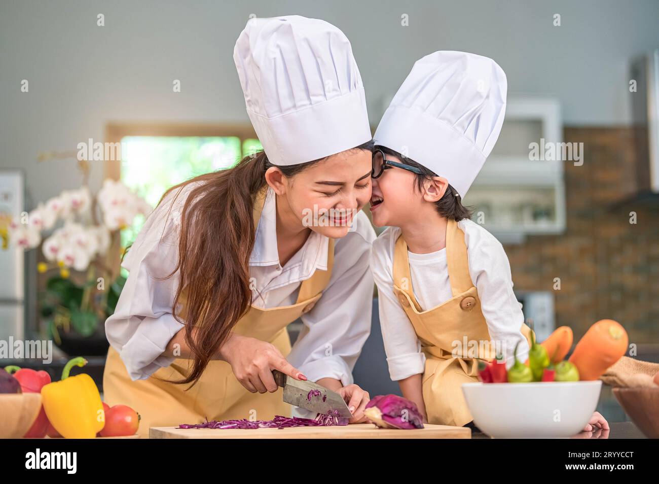 Happy cute little boy with eyeglasses kissing beautiful Asian woman mother while prepare to cooking in kitchen at home. People l Stock Photo