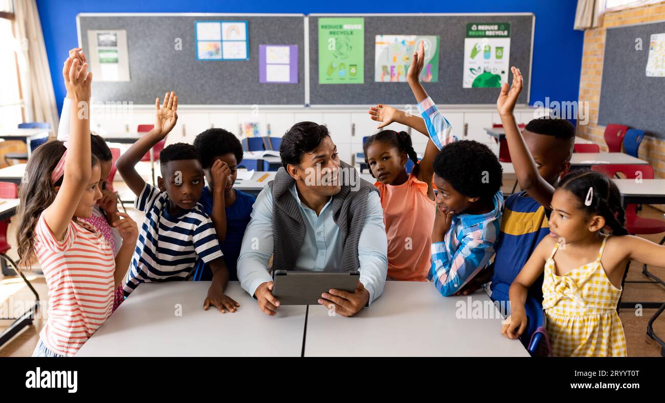 Diverse male teacher using tablet with children raising hands in class ...