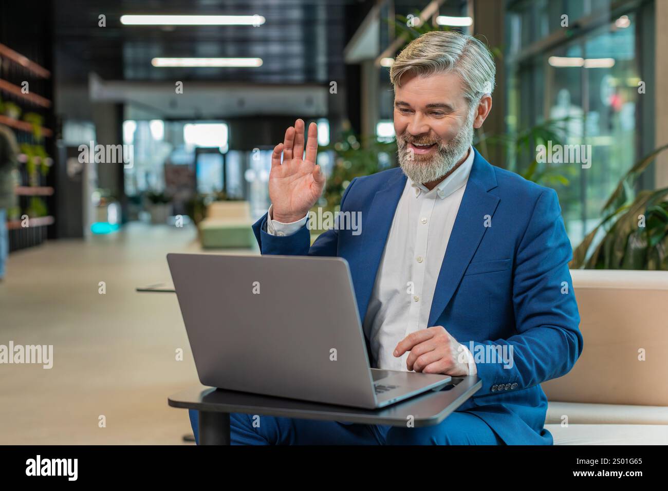 Middle-aged businessman working on laptop computer in modern office lobby talking on online communication video call with employee, boss. Manager man support services looking at webcam of computer. Stock Photo