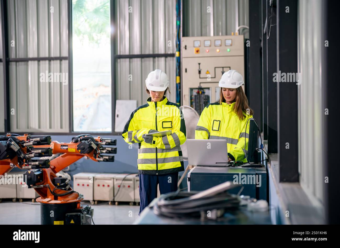Two workers wearing safety helmets and bright yellow jackets check data on a laptop while overseeing robotic arms in an industrial setting. The enviro Stock Photo