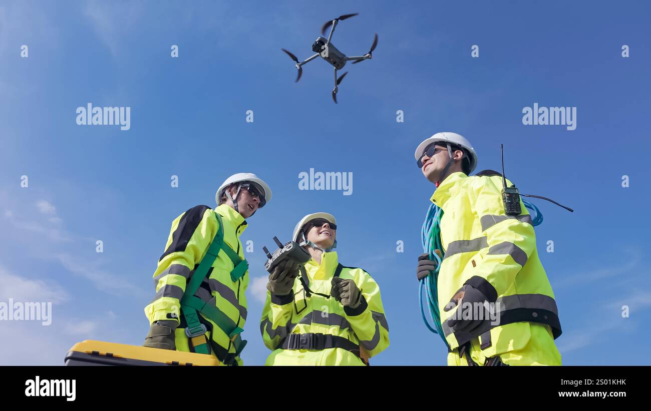 Three workers in high-visibility jackets monitor a drone in the sky while conducting aerial surveys at a construction site. The bright blue sky provid Stock Photo