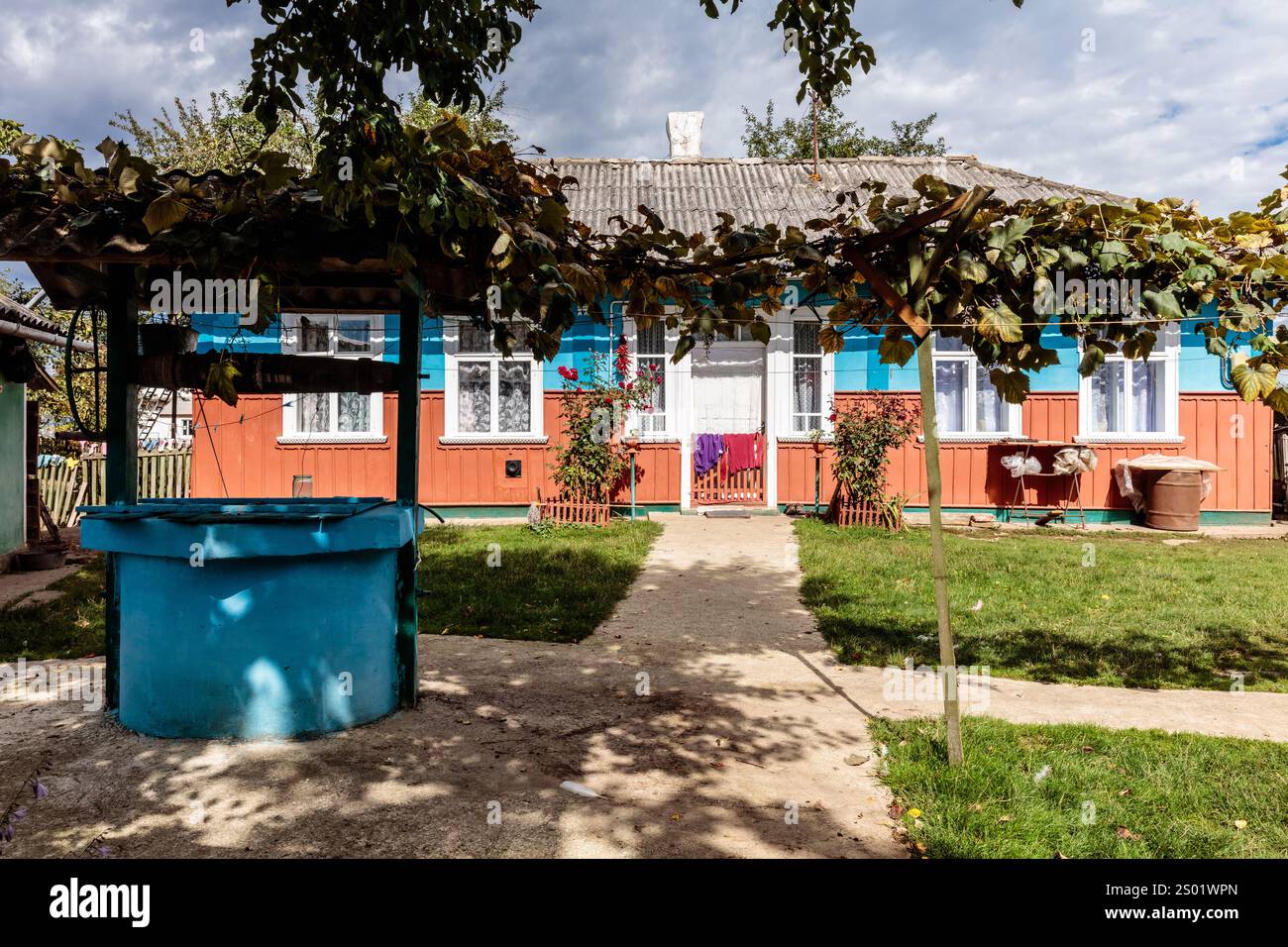 A blue house with a well in front of it. The house is surrounded by a garden with a lot of plants Stock Photo