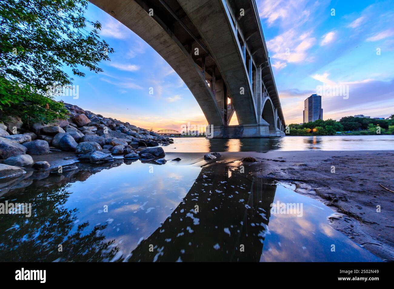 A bridge spans a river with a city in the background. The sky is a mix of blue and orange, creating a serene and peaceful atmosphere Stock Photo