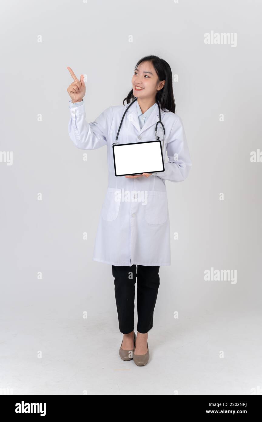 A full-body of a beautiful Asian female doctor in a white gown uniform showing a digital tablet with a blank screen to the camera, standing against an Stock Photo