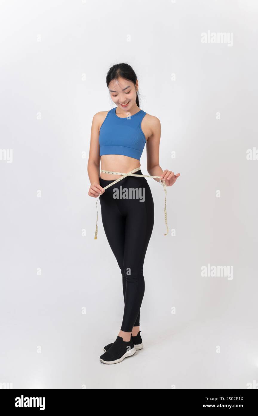 a-full-body-portrait-of-a-slim-healthy-asian-woman-in-sportswear-measuring-her-waist-with-a-measuring-tape-against-a-white-studio-background-dieting-2S02P1X.jpg