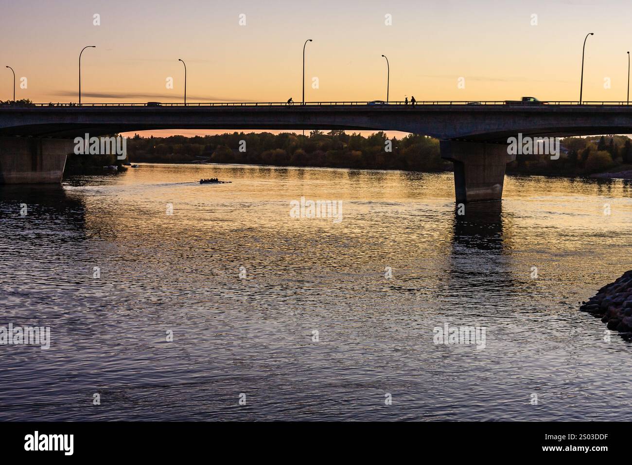 A bridge spans a river with a sunset in the background. The bridge is illuminated by street lights and the water is calm Stock Photo