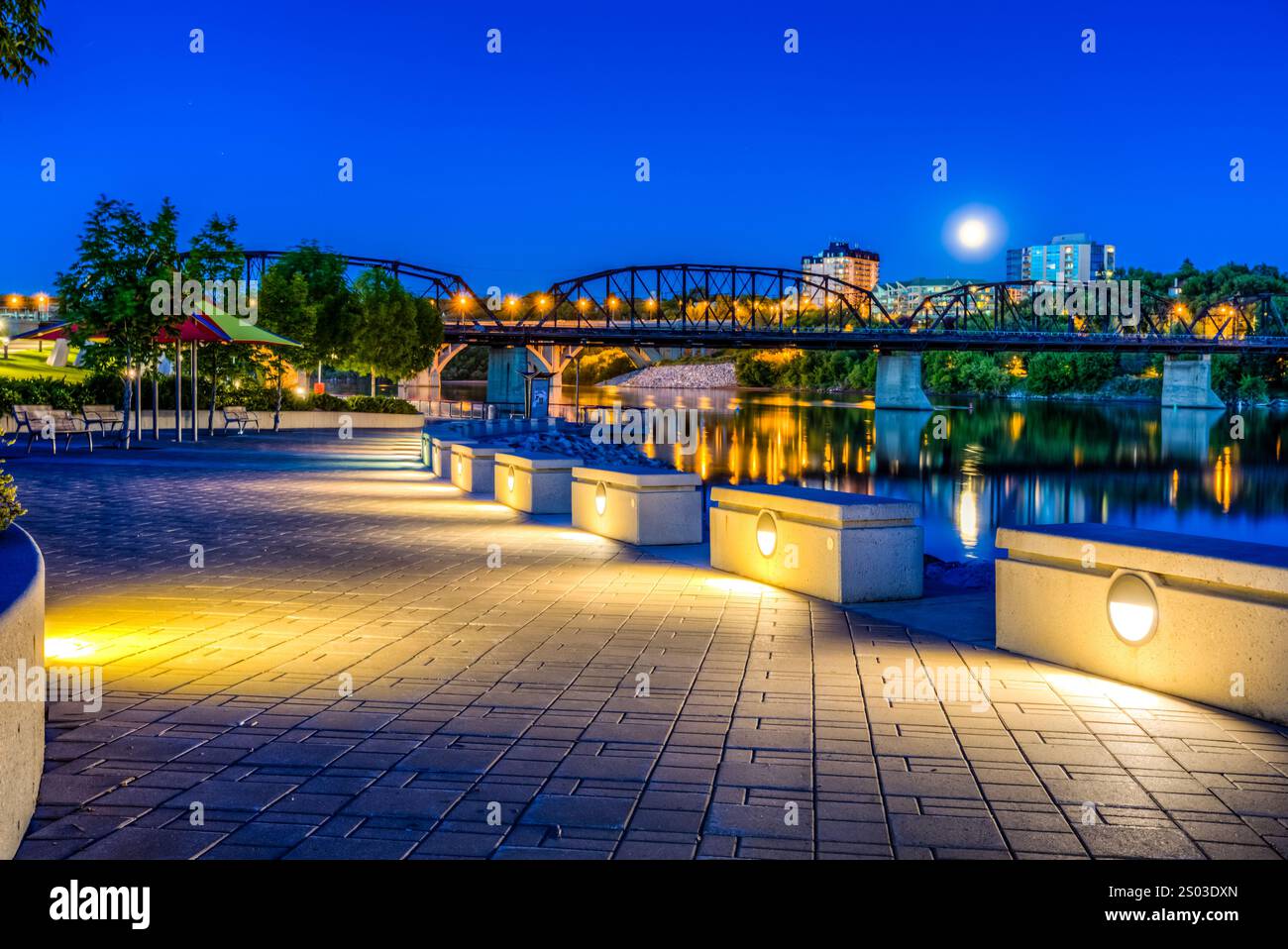 A city park with a bridge and a river. The bridge is lit up at night. The moon is visible in the sky Stock Photo