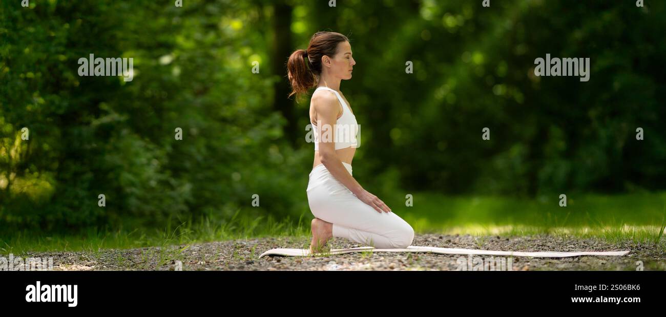 Woman kneeling on an exercise mat practicing yoga outdoors in the park Stock Photo