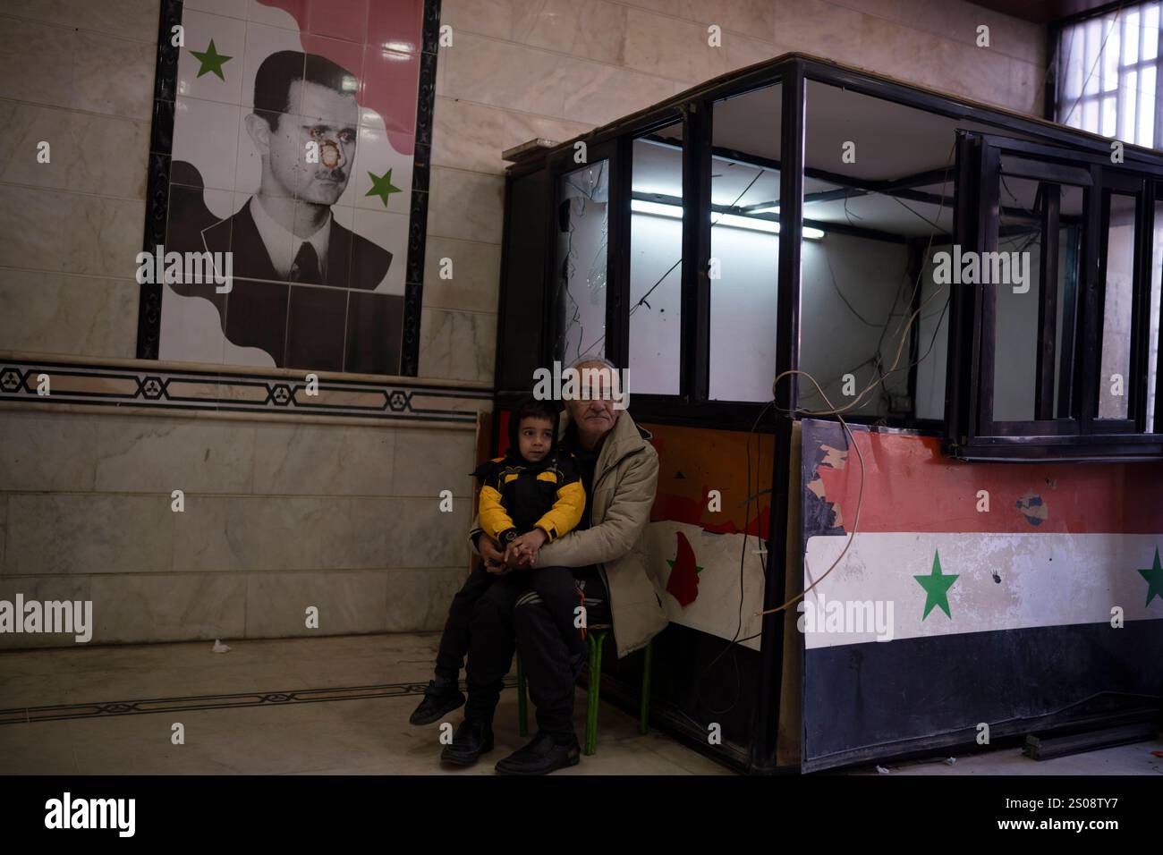 A man sits with a child next to a damaged image depicting the ousted ...