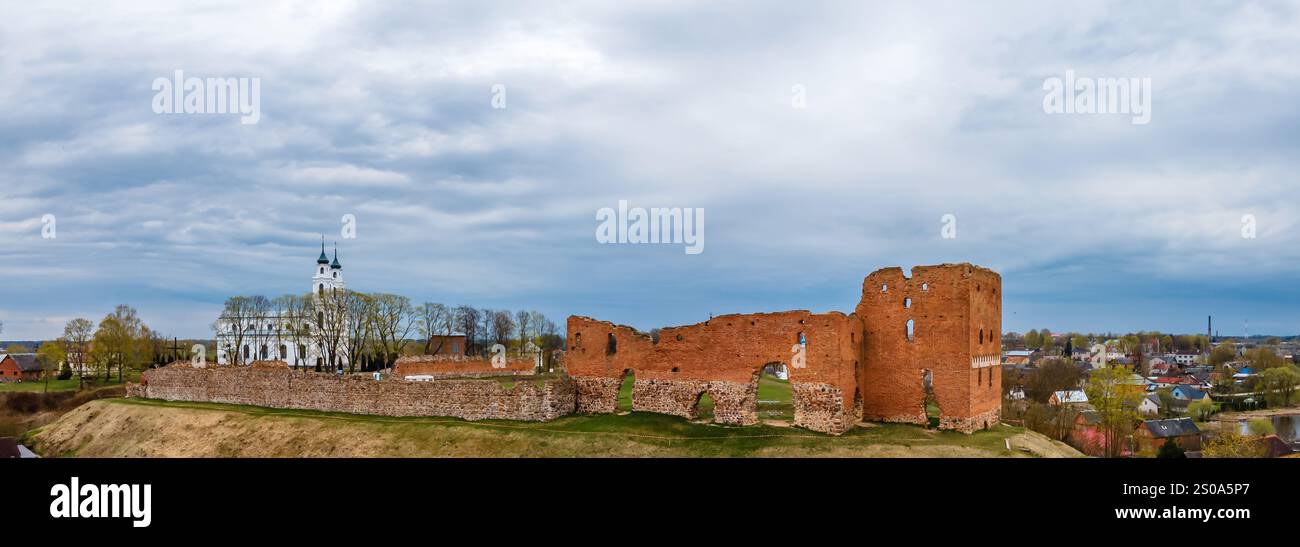 The image shows the ruins of a red brick castle with arched openings, set against a small town. A white church with twin spires is visible, surrounded Stock Photo