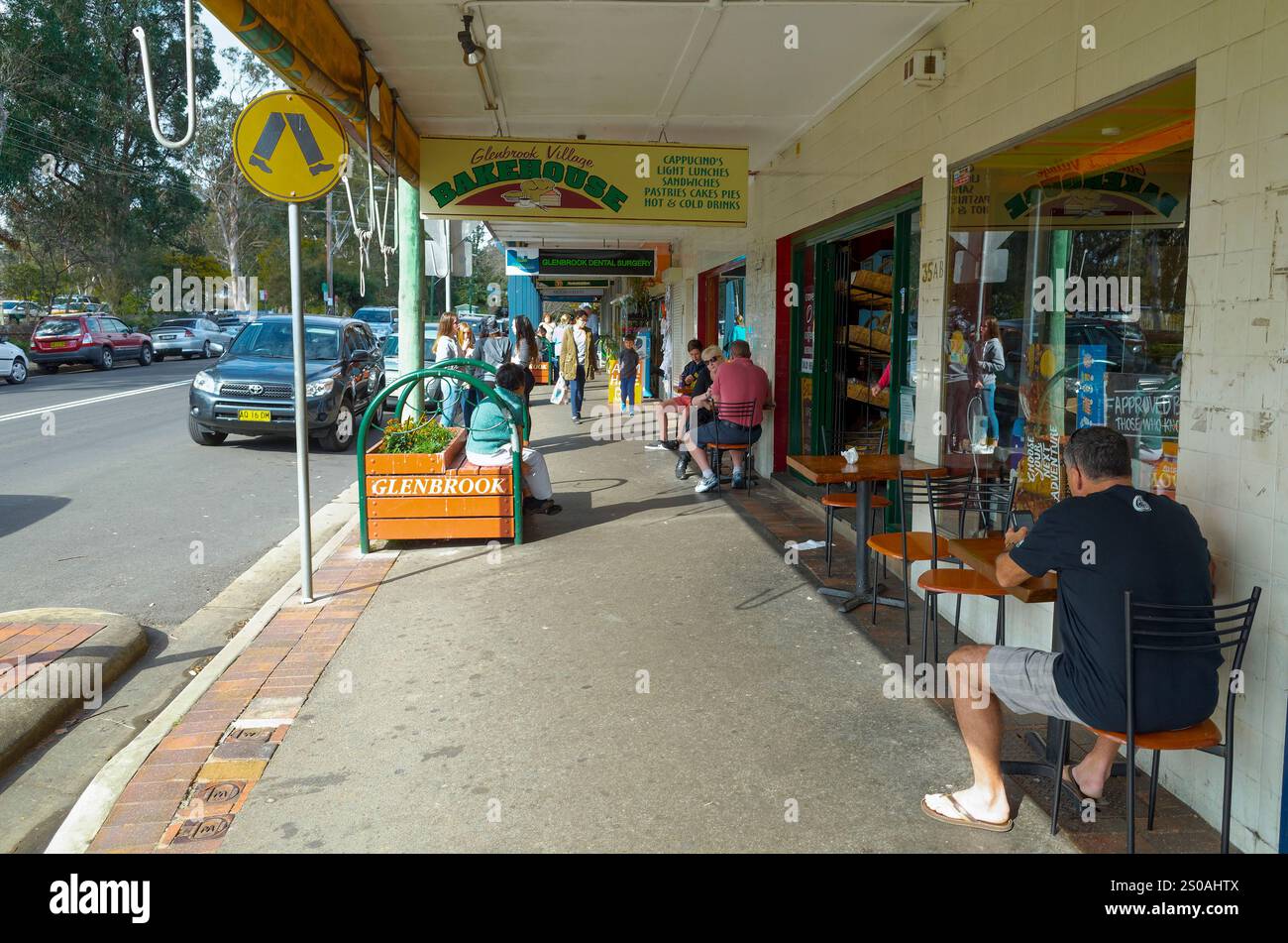 A bustling shopping strip in Glenbrook, Blue Mountains, with outdoor seating, locals enjoying the sunny atmosphere, parked cars, and shops like Glenbr Stock Photo