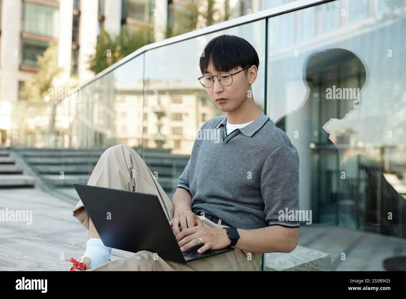 Medium shot of Asian male IT programmer typing on laptop sitting on sidewalk leaning on glass construction while working in public space in city street, copy space Stock Photo