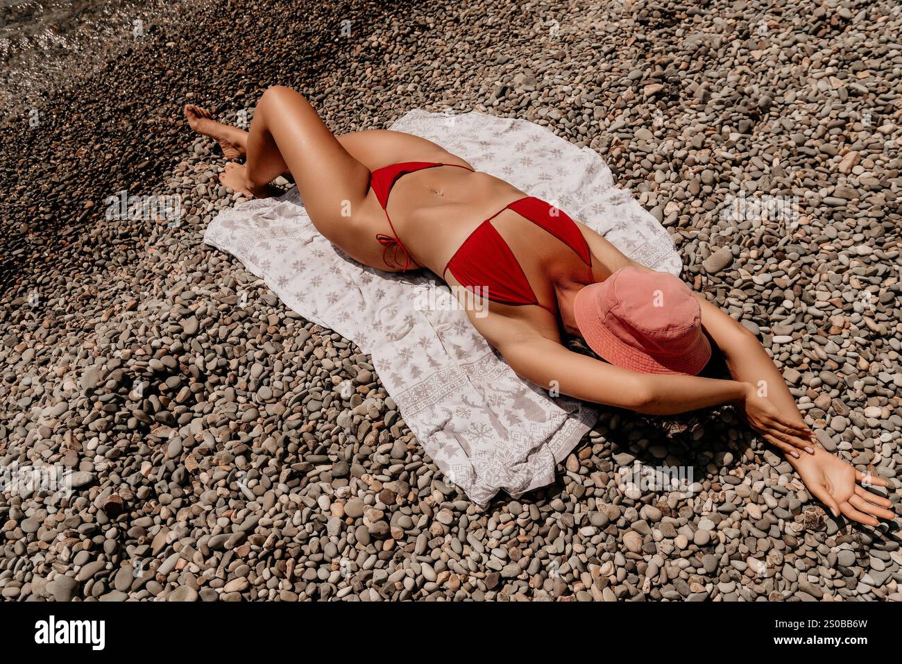 Woman Sunbathing Beach Summer - A woman in a red bikini is sunbathing on a beach. She is lying on a towel and wearing a pink hat. The scene is sunny Stock Photo