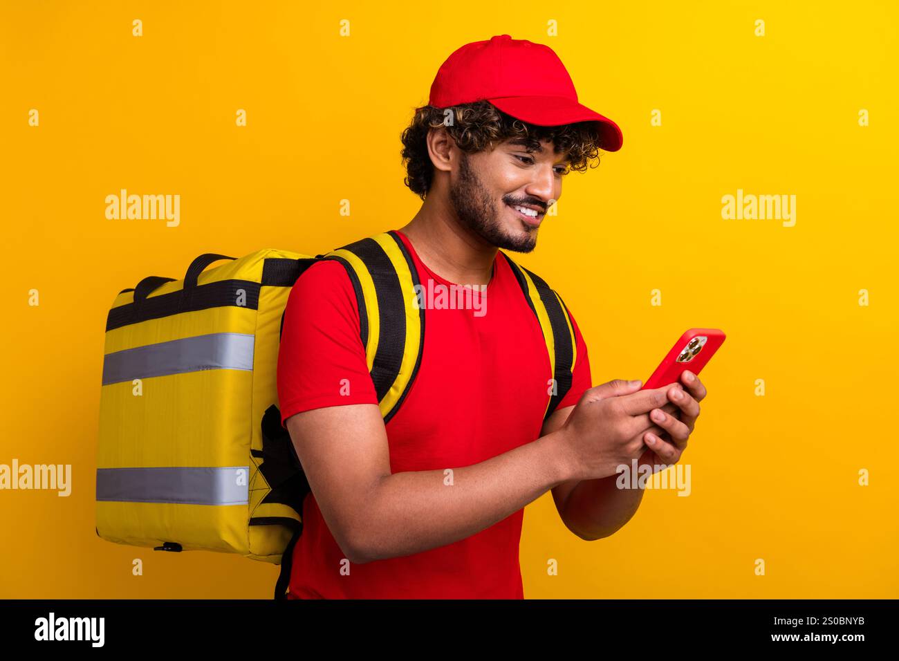 Cheerful courier with yellow backpack using smartphone on a vibrant colored background. Stock Photo