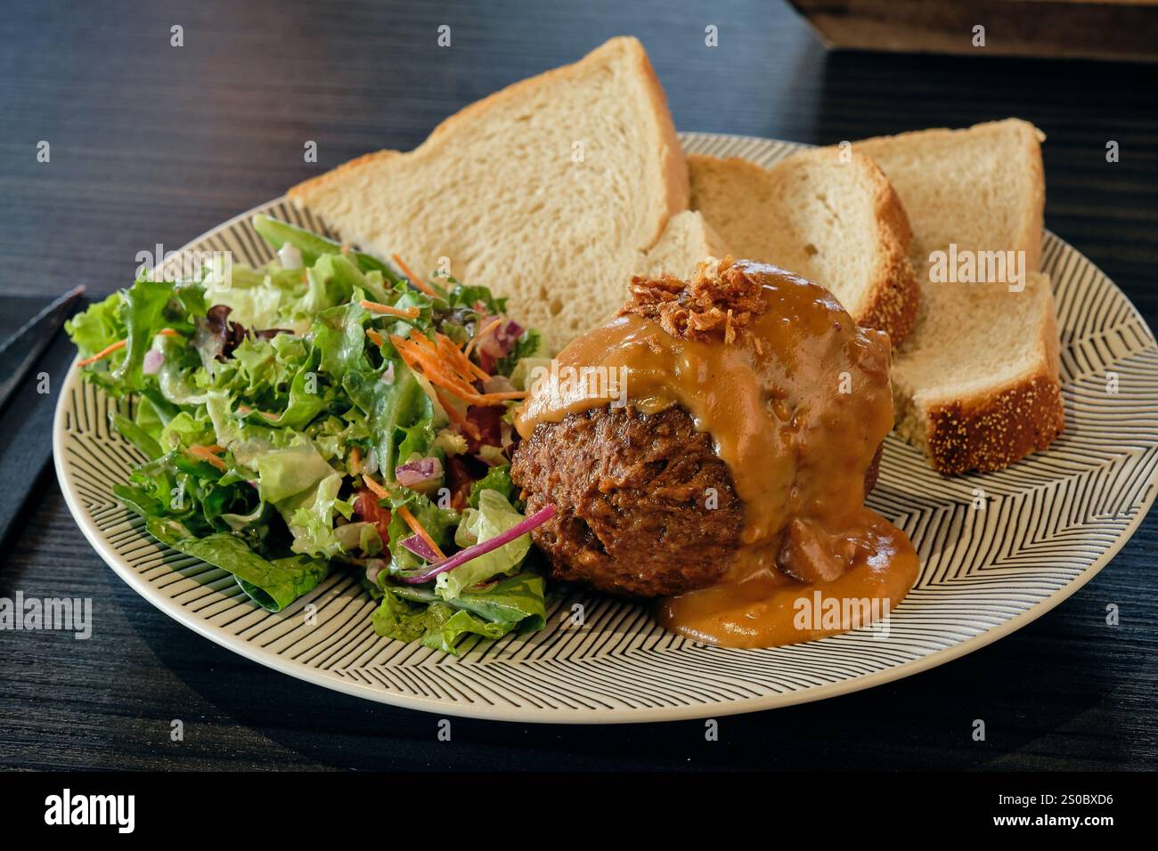 A flavourful Dutch dish featuring a meatball topped with satay sauce, served with fresh salad and slices of white bread on a patterned plate. Stock Photo