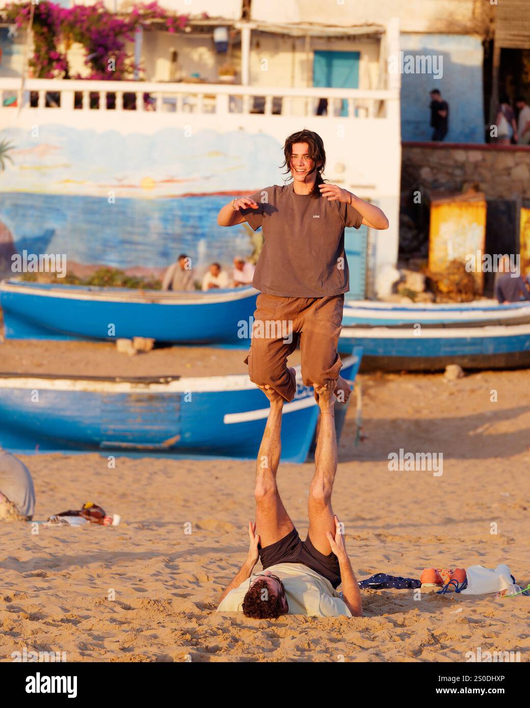 Young woman smiles whilst doing a balancing act with a friend in front of the blue fishing boats on the beach in Taghazout, Morocco. Dec 27, 2024. Stock Photo