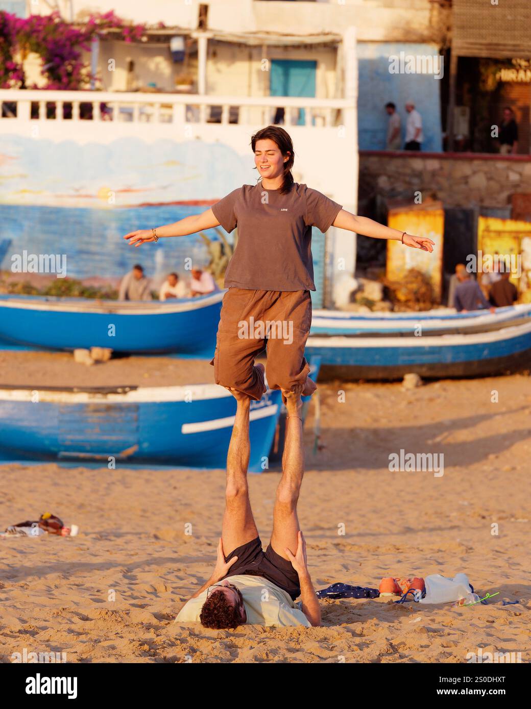 Young woman smiles whilst doing a balancing act with a friend in front of the blue fishing boats on the beach in Taghazout, Morocco. Dec 27, 2024. Stock Photo