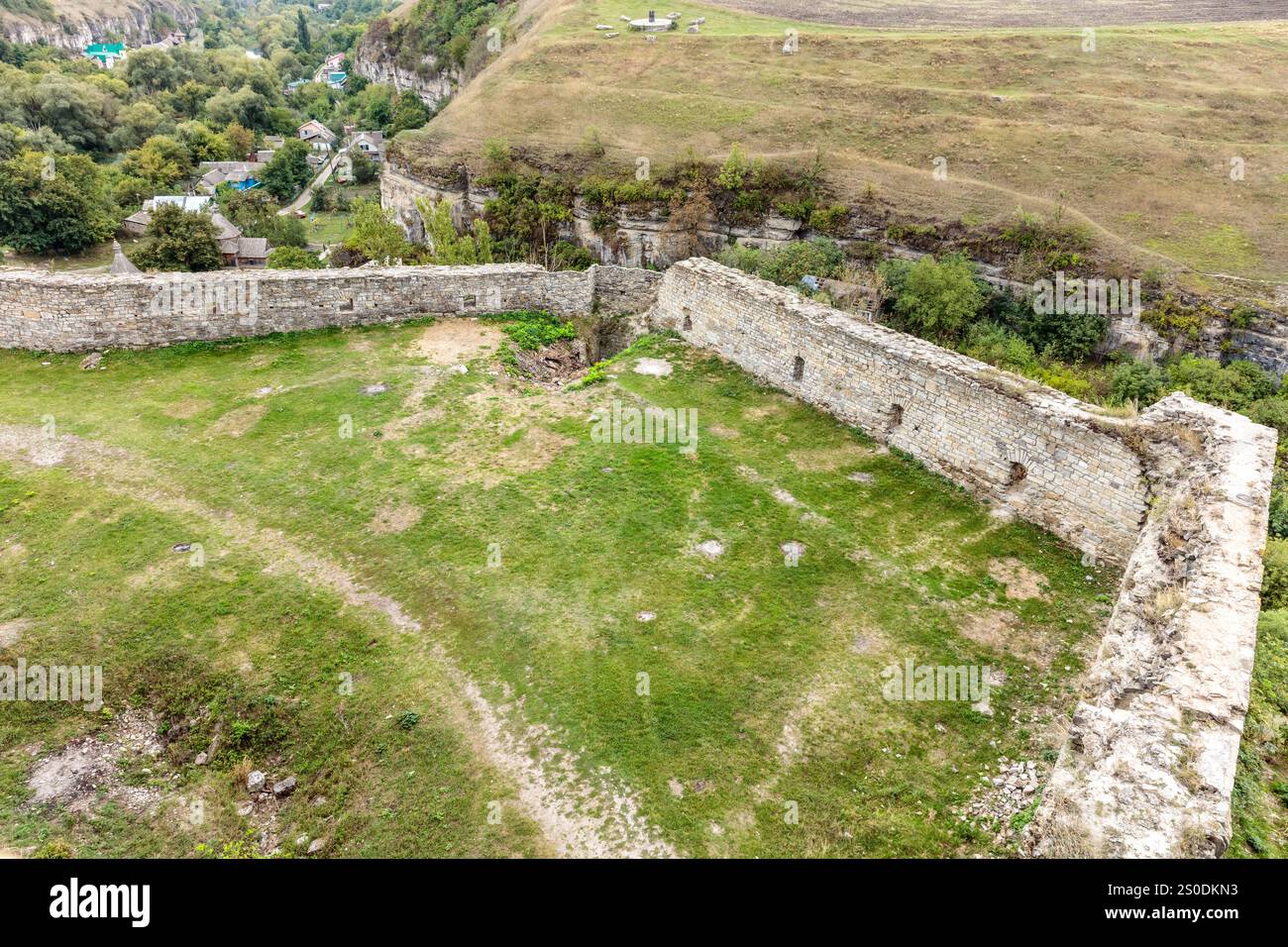 A large stone wall with a grassy field in front of it. The wall is old and has a lot of cracks. The grass is green and well-maintained Stock Photo