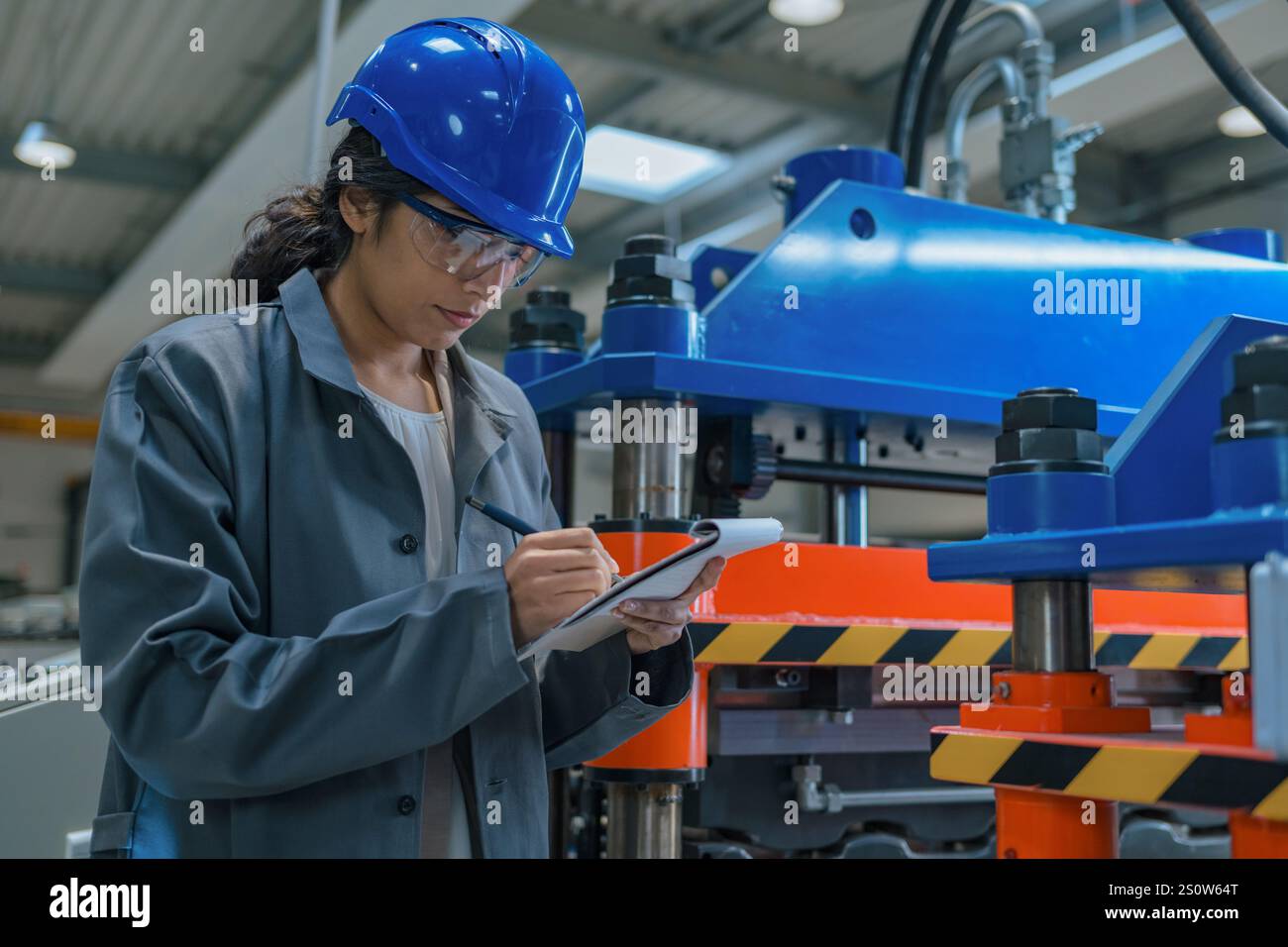 Indian woman engineer working in a production line, examining automated machinery work and writing down a planning checklist. Stock Photo