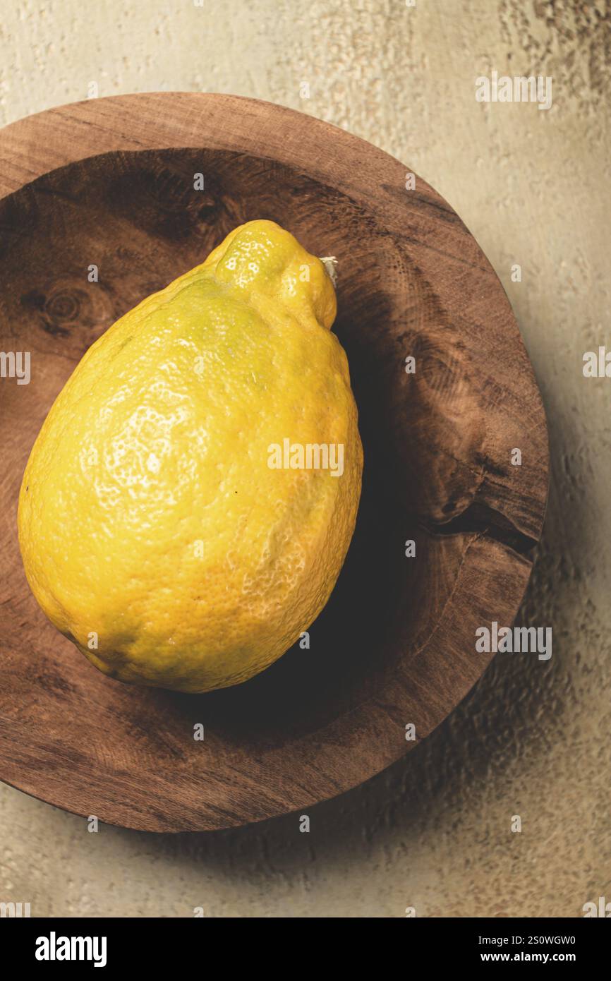 A vibrant yellow lemon sits atop a rustic wooden plate, showcasing its bumpy texture and bright color against a softly illuminated background, highlig Stock Photo