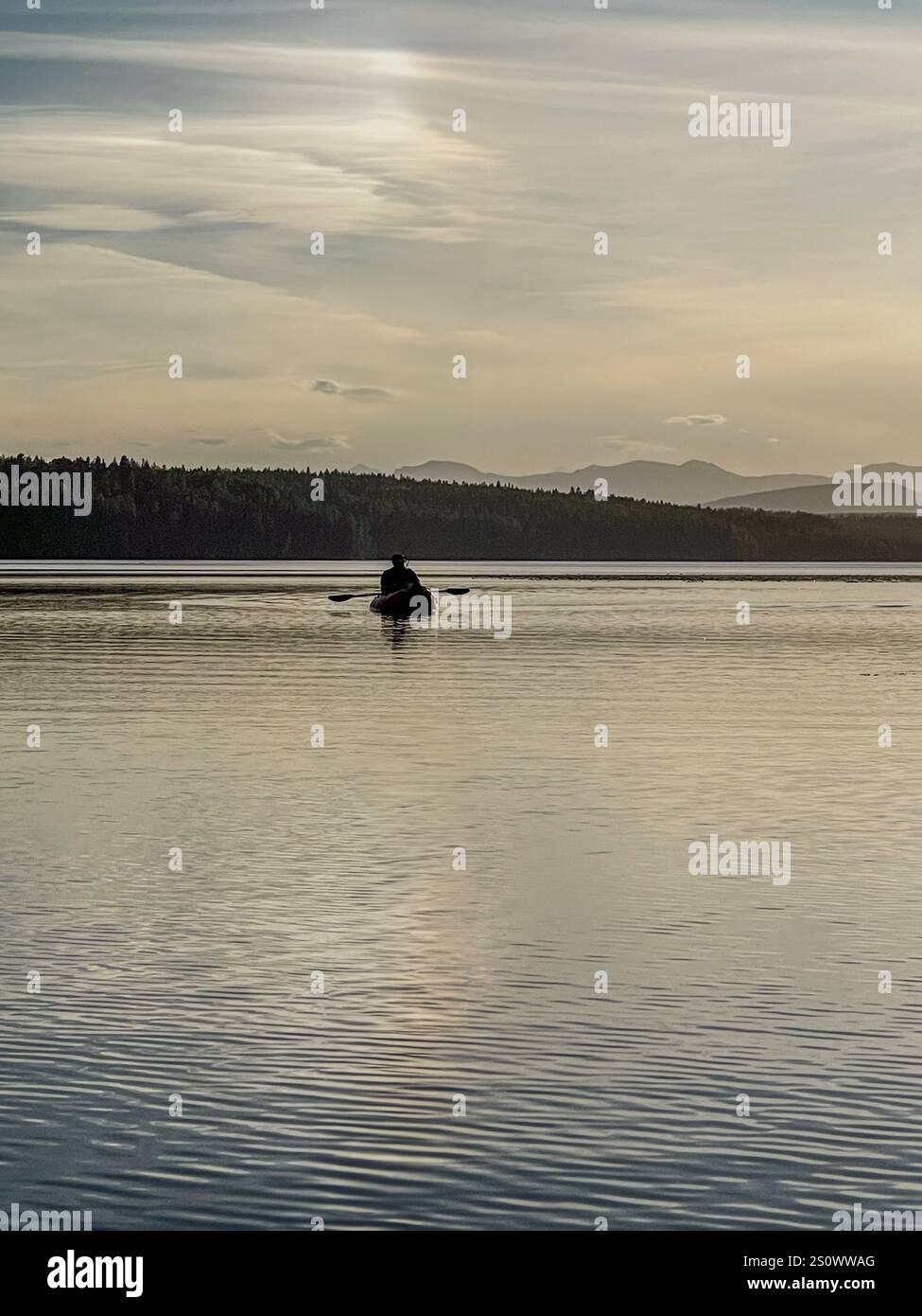 A single kayaker paddling on a calm lake at sunset, surrounded by soft mountain silhouettes and clouds. - Smartphone Captured Stock Image