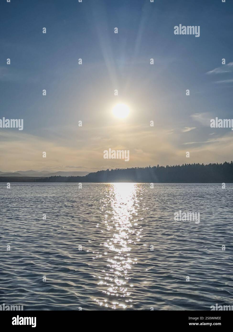 Scenic view of a serene lake with sunlight reflecting on the water during dusk, framed by distant forested hills and a clear blue sky. - Smartphone Captured Stock Image