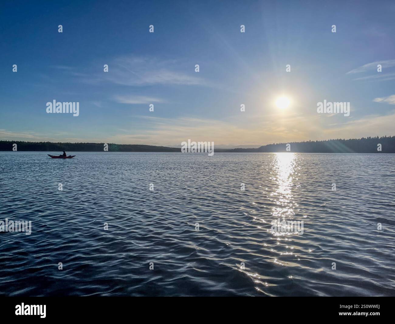 A lone kayaker paddles across a serene lake under a soft sunset, surrounded by distant forested hills and a calm atmosphere. - Smartphone Captured Stock Image