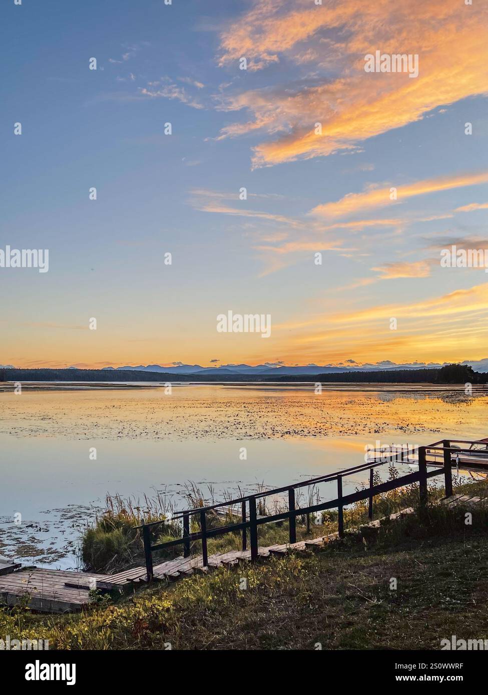 A scenic sunset over a tranquil lake with a wooden dock and railings in the foreground. - Smartphone Captured Stock Image