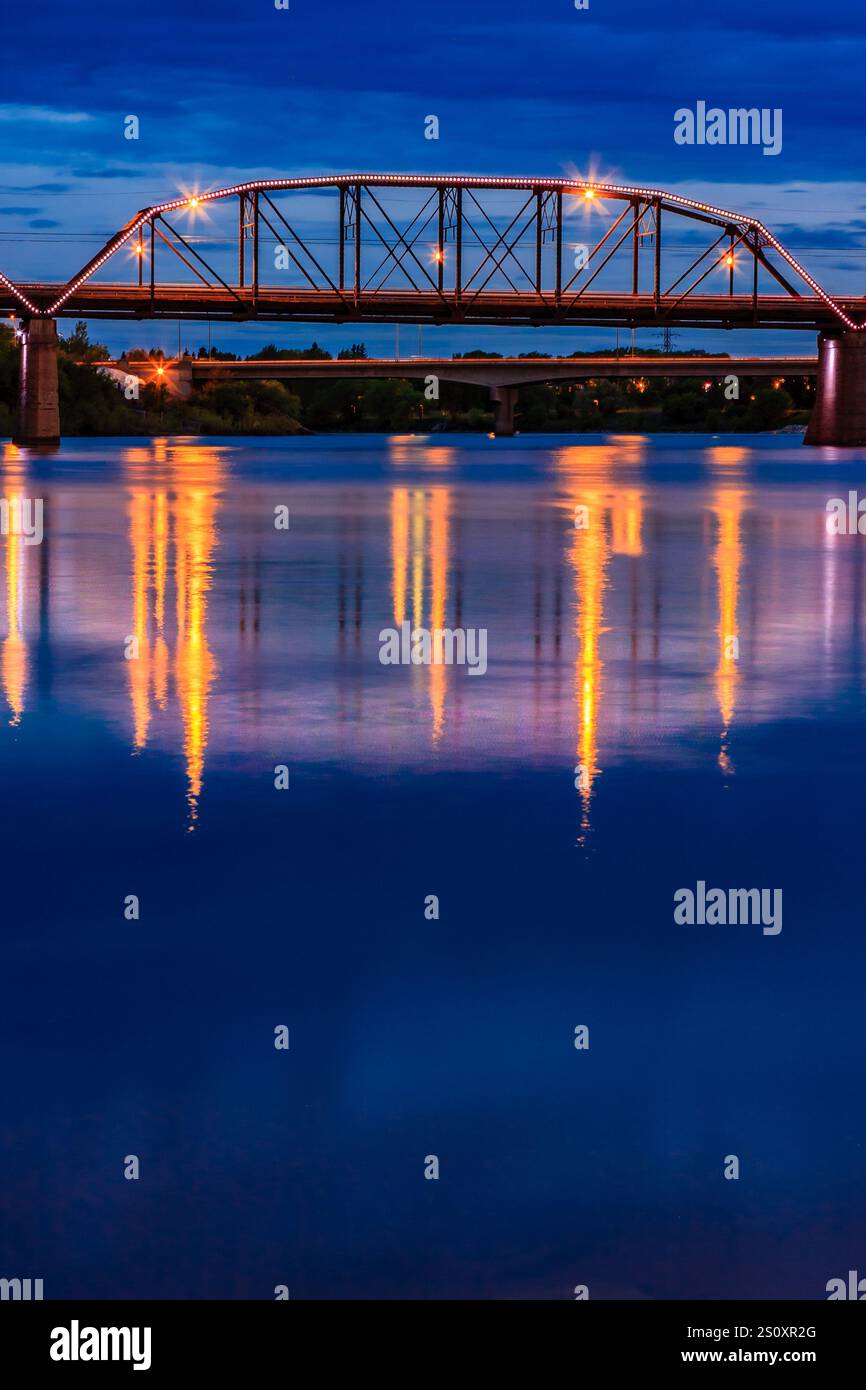A bridge spans a river at night. The bridge is lit up with lights, and the water is calm Stock Photo