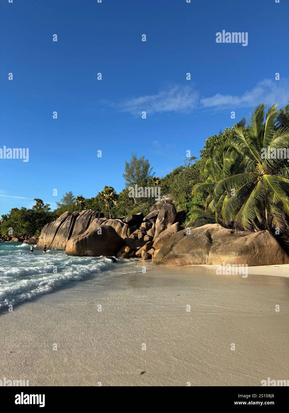 A secluded tropical beach with smooth granite boulders, lush green palm trees, and crystal-clear waters lapping at the white sandy shore - Smartphone Captured Stock Image