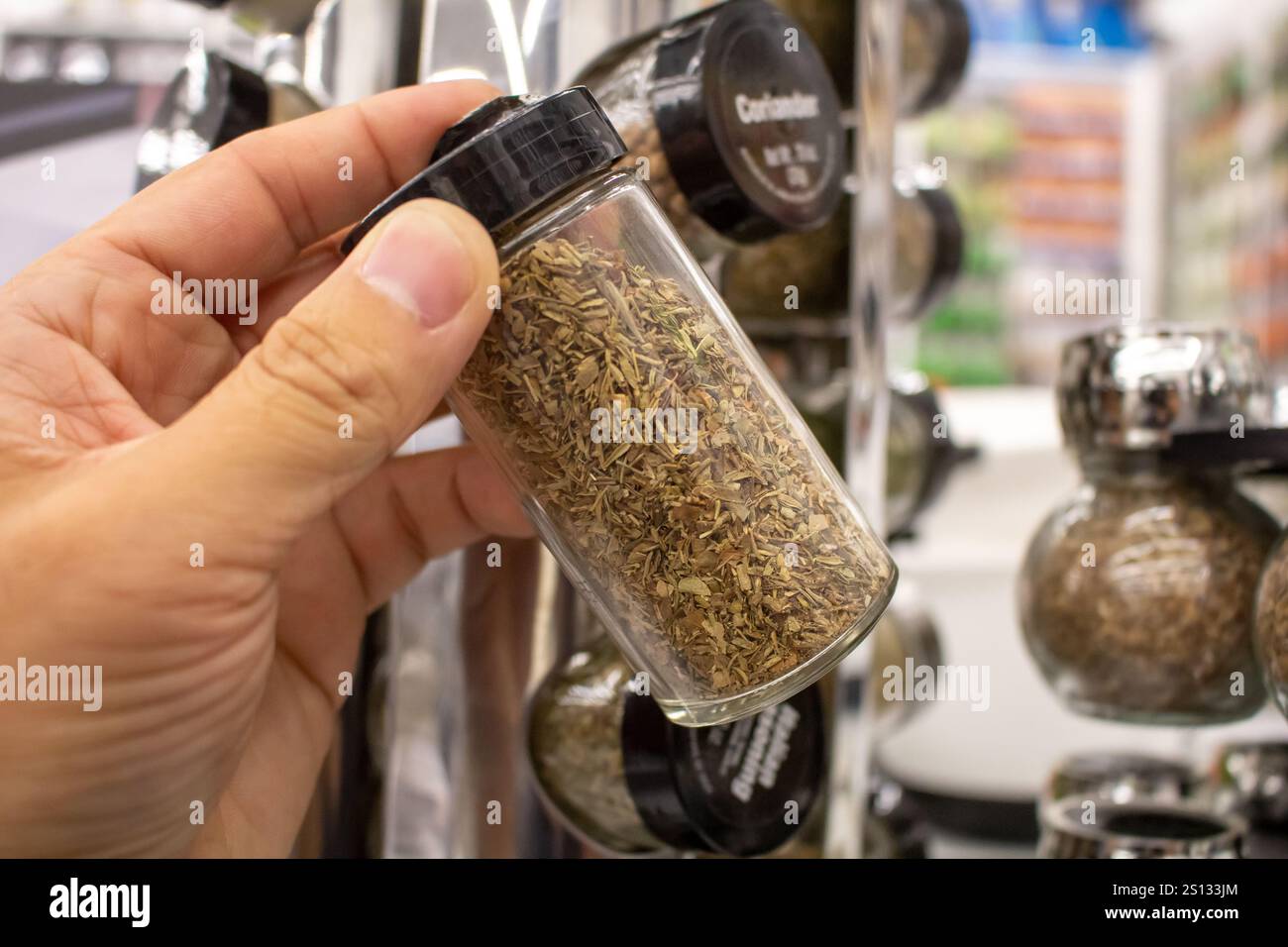A view of a hand holding a spice jar in front of several other spice jars on a spice rack, at the store. Stock Photo