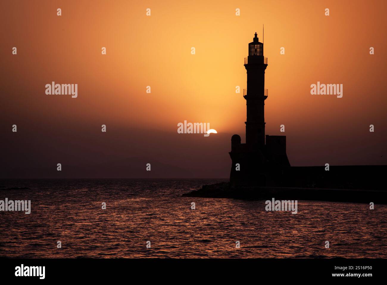 The Chania lighthouse stands proudly at the entrance of the Venetian harbor in Chania, Crete, casting its silhouette against the shimmering waters. Stock Photo