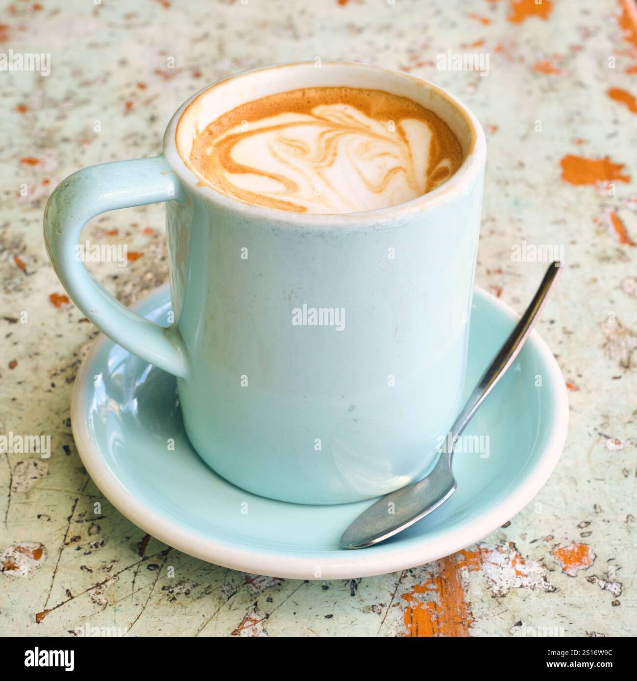 A flat white coffee in a pale blue mug and saucer with teaspoon on a worn, rustic tabletop that matches in hues of pale blue and orange. Stock Photo